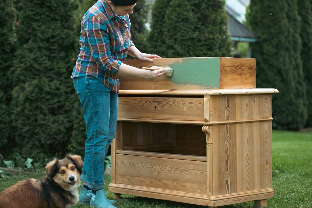 Woman painting a wooden furniture while sitting with her dog in the garden. DIY concept.