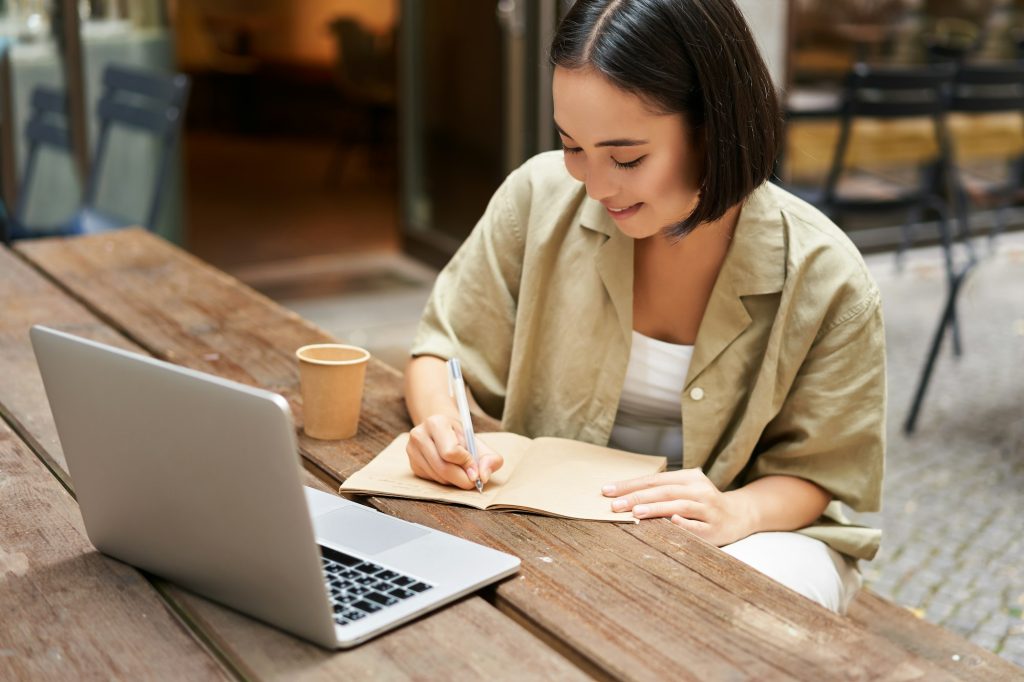 Portrait of young asian woman working on laptop, making notes, writing down while attending online